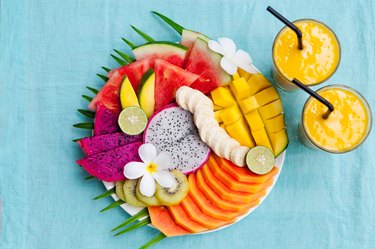 Assortment of fruits that contain serotonin on a white plate with palm tree leaf with mango smoothie, textile blue background. Top view. Copy space