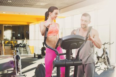 Fitness instructor helps young woman on elliptical trainer