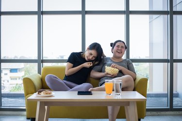 Overweight woman and asian girl enjoy eating food on sofa at home