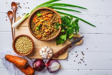 Lentil soup in a wooden bowl and ingredients on a white wooden background, top view.