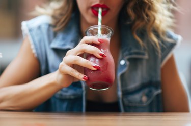 close view of a person wearing a jean vest drinking cranberry juice from a glass with a blue-and-white striped straw