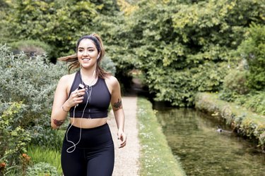 Young female runner listening to earphones while running on riverside path