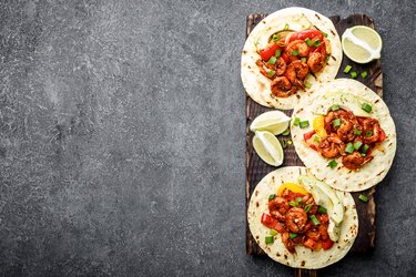 Fajitas in tortillas with fried shrimps, bell peppers and onion served up with avocado and green onions on wooden cutting board, top view, food background with space for a text