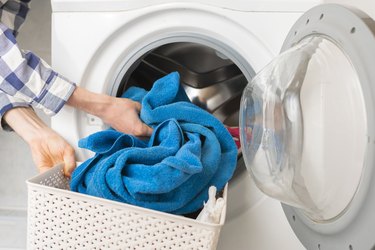 a close up of a person's hands putting a blue towel in a washing machine from a hamper