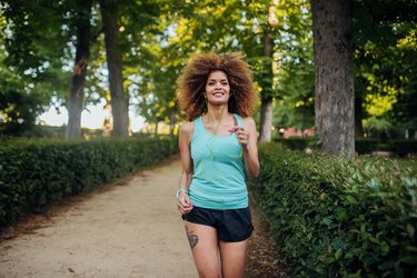 A jogger with natural hair runs in a park surrounded by trees and bushes