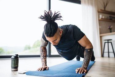 A fit young adult with dreadlocks doing a push-up on a blue yoga mat at home