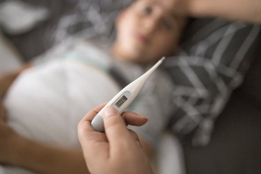 foreground view of a thermometer registering a 100 degree fever, with a blurred child in the background