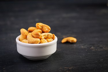 a small white bowl of cashews with one out of the bowl on dark wooden background