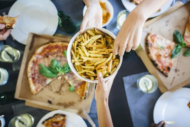 overhead photo of hand taking french fries from bowl with pizza in background