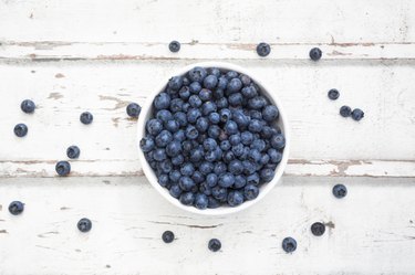 Top-down photograph of a white bowl full of blueberries on a white wooden table with some blueberries scattered lose around the bowl