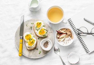 Morning breakfast table inspiration - sandwiches with cream cheese and boiled egg, yogurt with apple and flax seeds, herbal detox tea, notebook, glasses on light background, top view. Flat lay