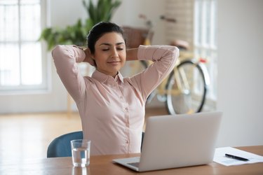 an adult wearing a light pink button down shirt relaxes with closed eyes and hands behind their head doing breathing exercises in front of a laptop