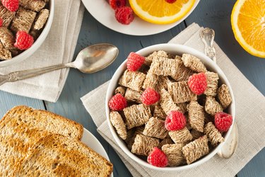 Healthy shredded wheat cereal in a white bowl with raspberries on a table.