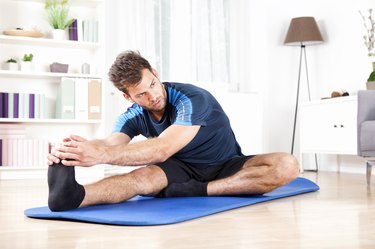 man doing a hamstring stretch sitting on blue yoga mat at home