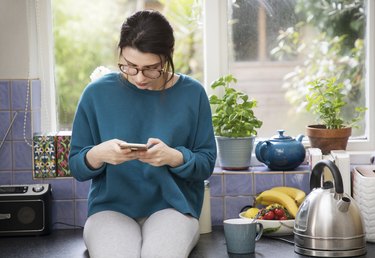 Woman using weight-loss incentive app on her phone, while sitting on the kitchen counter next to a tea kettle