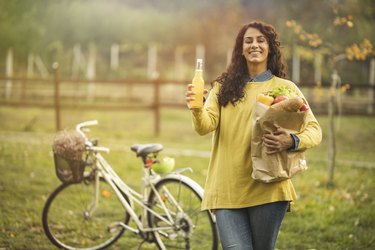 Woman drinking juice and holding shopping bag near her bicycle
