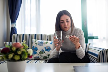 a person wearing a gray turtleneck sits on a striped couch and reads the back of a package of pills holding a glass of water