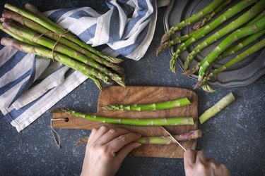 Fresh raw asparagus on stone background. Female hands cut asparagus on wooden cooking board. Top view