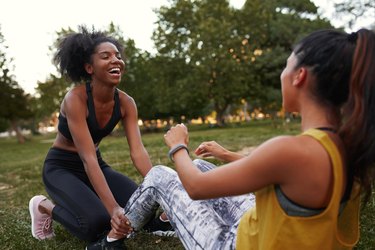 Cheerful young diverse female friends laughing doing ab crunches on green grass in the park - friends laughing together while doing exercise outdoors