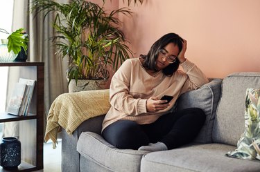 lonely woman in peach sweater sitting on gray couch looking down at smartphone