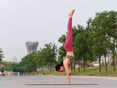 Side view of healthy women in sportswear, red pants and top practicing yoga outdoor, doing handstand exercise on yoga mat salamba sirsasana pose, full length portrait.