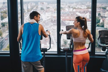 Back view of smiling athletic couple talking while exercising on cross trainer in a gym.