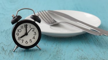 Intermittent fasting concept showing an alarm clock next to an empty plate with silverware on a blue wooden table