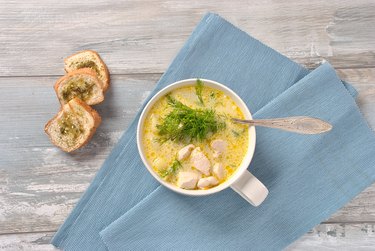 an overhead photo of a mug of chicken soup with spices herbs and seasonings on a blue cloth napkin on top of a wooden table