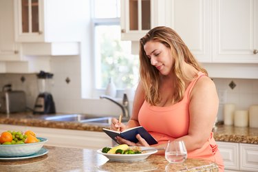 Person in their kitchen writing down their caloric intake while eating breakfast.