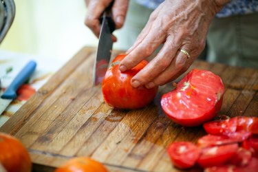 Close-up of Senior Adult Woman Slicing Tomato on Wooden Cutting Board - Stock Photo