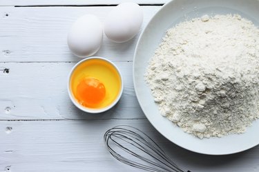 A plate with flour, eggs, a whisk on a white wooden background. Pancakes ingredients. Egg in the bowl.
