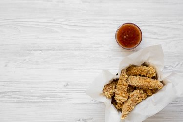 Chicken fingers in paper box with sauce on a white wooden background.