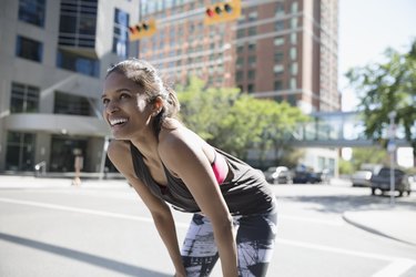 Smiling female runner resting with hands on knees on sunny urban street