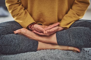 Close up of a person wearing gray leggings and a yellow long-sleeve shirt sitting crosslegged meditating indoors