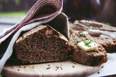 Loaf of freshly baked homemade whole grain rye bread with seeds cut in two on a wooden cutting board and covered with linen tea towel.