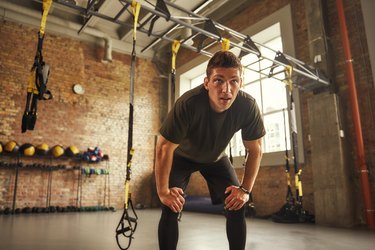 an athlete in a gym with their hands on their knees looking tired during a workout at a gym with TRX straps in the background