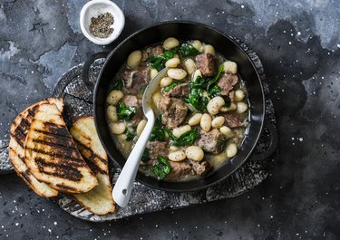 Beef, beans, spinach slow cooker stew in a pan on a wooden board on a dark background. Delicious homemade comfort food