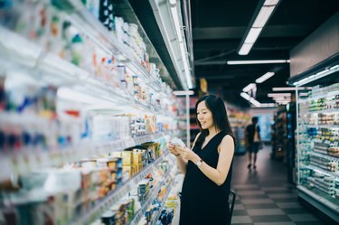 Asian pregnant woman grocery shopping in supermarket and reading nutrition label on a packet of cheese