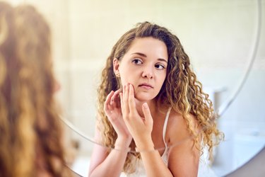 A woman with curly hair looking in the mirror at acne caused by too much added sugar
