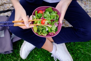 A girl sits on the grass and eats a green fresh salad with chopsticks for sushi.