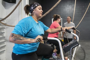 Two attractive body-positive women, Latino and Caucasian, doing a workout on the exercise bike in the gym under the supervision of the coach, the senior 55-years-old Cuban Hispanic man