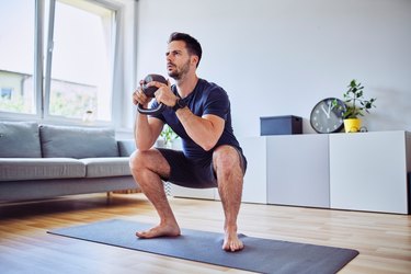 Training at home, young man doing squats with kettlebell weights.