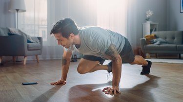 Strong Athletic Fit Man in T-shirt and Shorts is Doing Mountain Climber Exercises While Using a Stopwatch on His Phone. He is Training at Home in His Spacious Apartment with Minimalistic Interior.