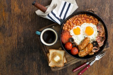 Full English breakfast with sausages, bacon, fried eggs, beans, toast and cup of coffee on wooden table with copy space