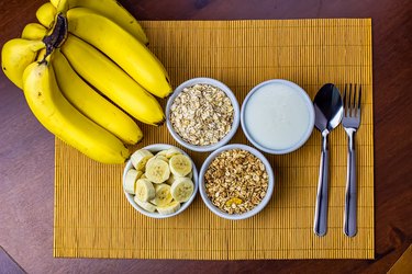 Banana sliced ramekin with oatmeal, granola and plain yogurt as side dishes under bamboo mat with bunch of bananas besides in top view