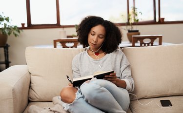 a person wearing a gray sweater sits on a tan couch at home writing in a gratitude journal