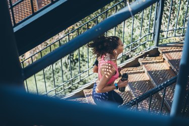 person with long curly hair in a ponytail and salmon-colored top running up outdoor stairs and holding a black water bottle