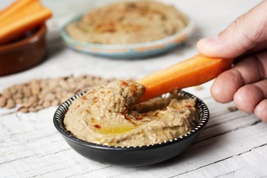 Person dipping carrot in a homemade lentil hummus in small black bowl on wooden table.