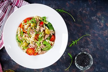 Pasta salad with tuna, tomatoes, olives, cucumber, sweet pepper and arugula on rustic background . Top view