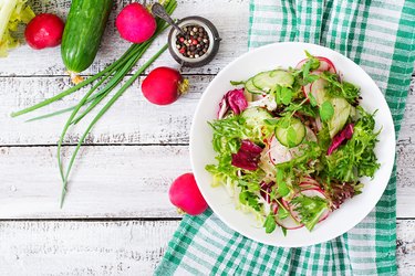 Top view of a bowl of fresh salad with cucumbers, radishes and herbs on a white wooden table.